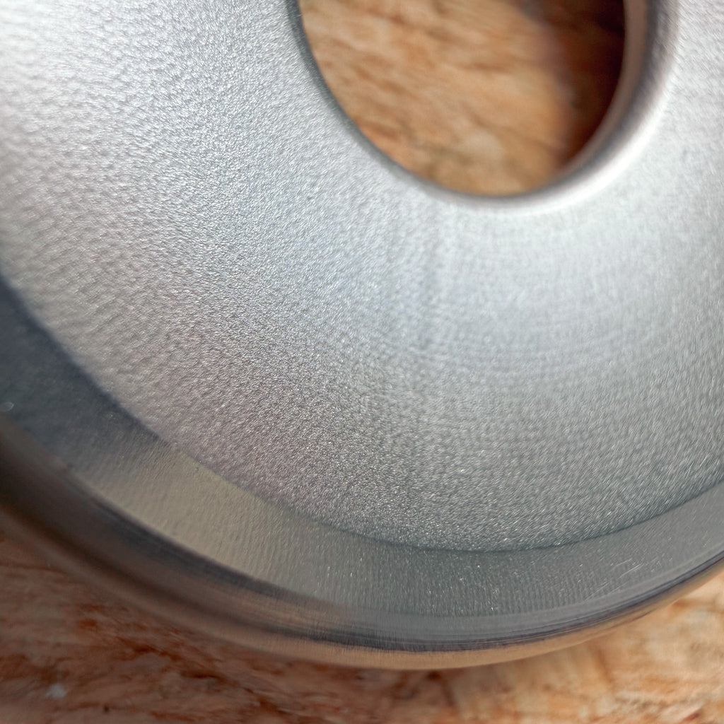 Close up of a circular metallic 316 stainless steel ring or coin with a sloping mildly textured surface on a textured plywood or OSB surface.