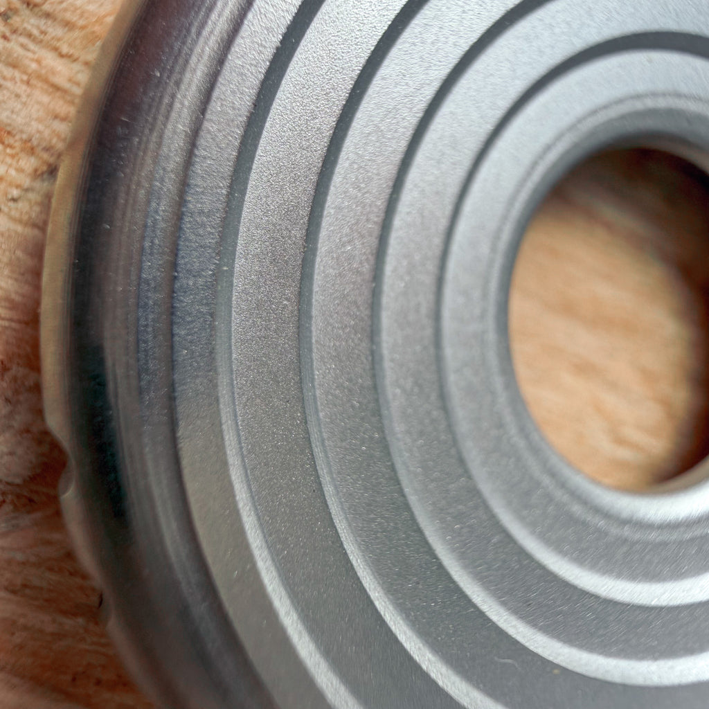 Close up of a circular metallic 316 stainless steel ring or coin with concentric grooves on a textured plywood or OSB surface.