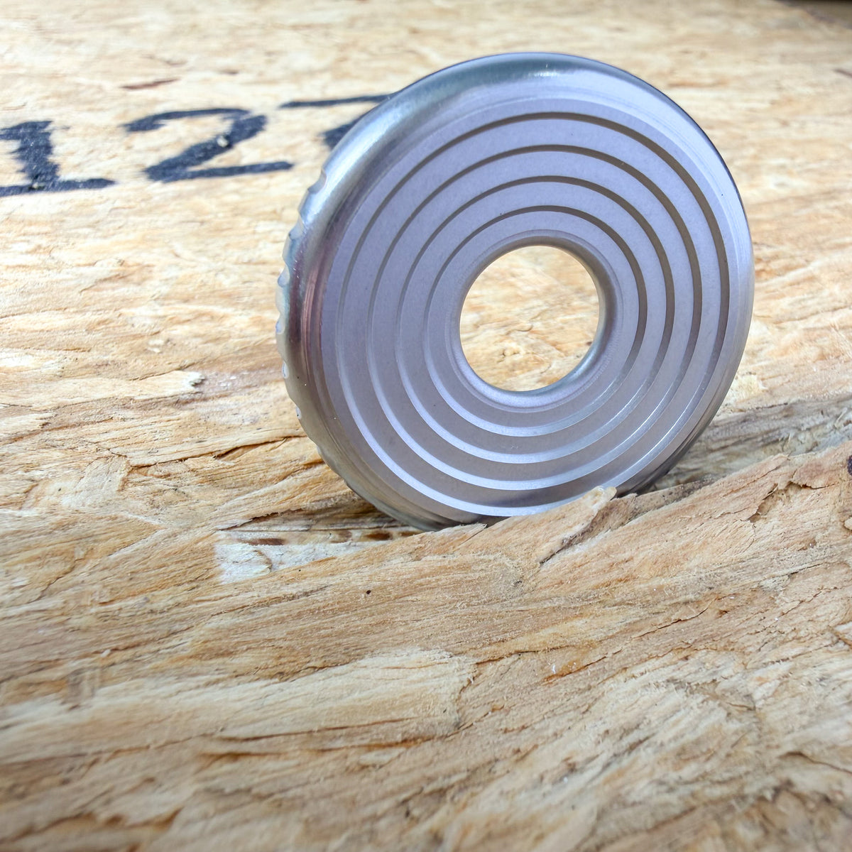 A circular metallic 316 stainless steel ring or coin with concentric grooves, standing upright on a textured plywood or OSB surface.