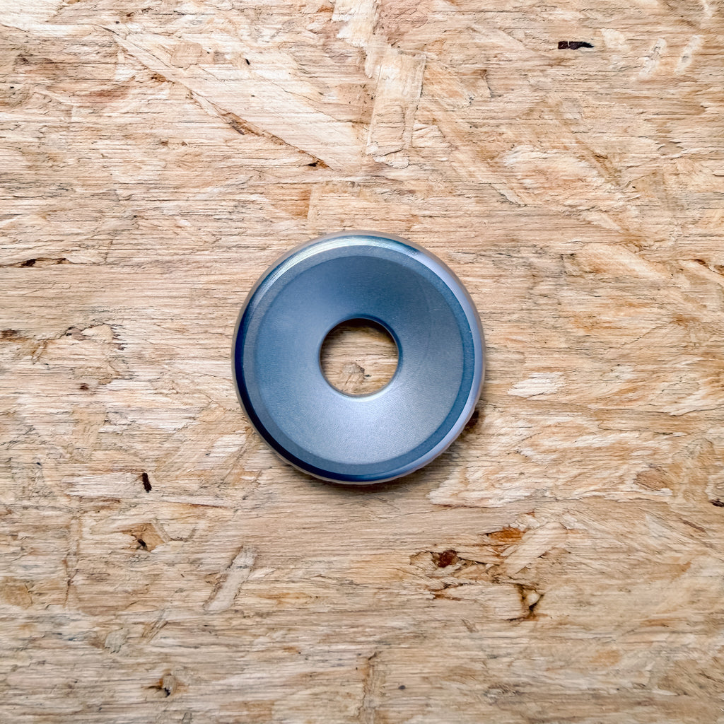 A circular metallic 316 stainless steel ring or coin with a smooth surface, placed flat on a textured plywood or OSB surface.