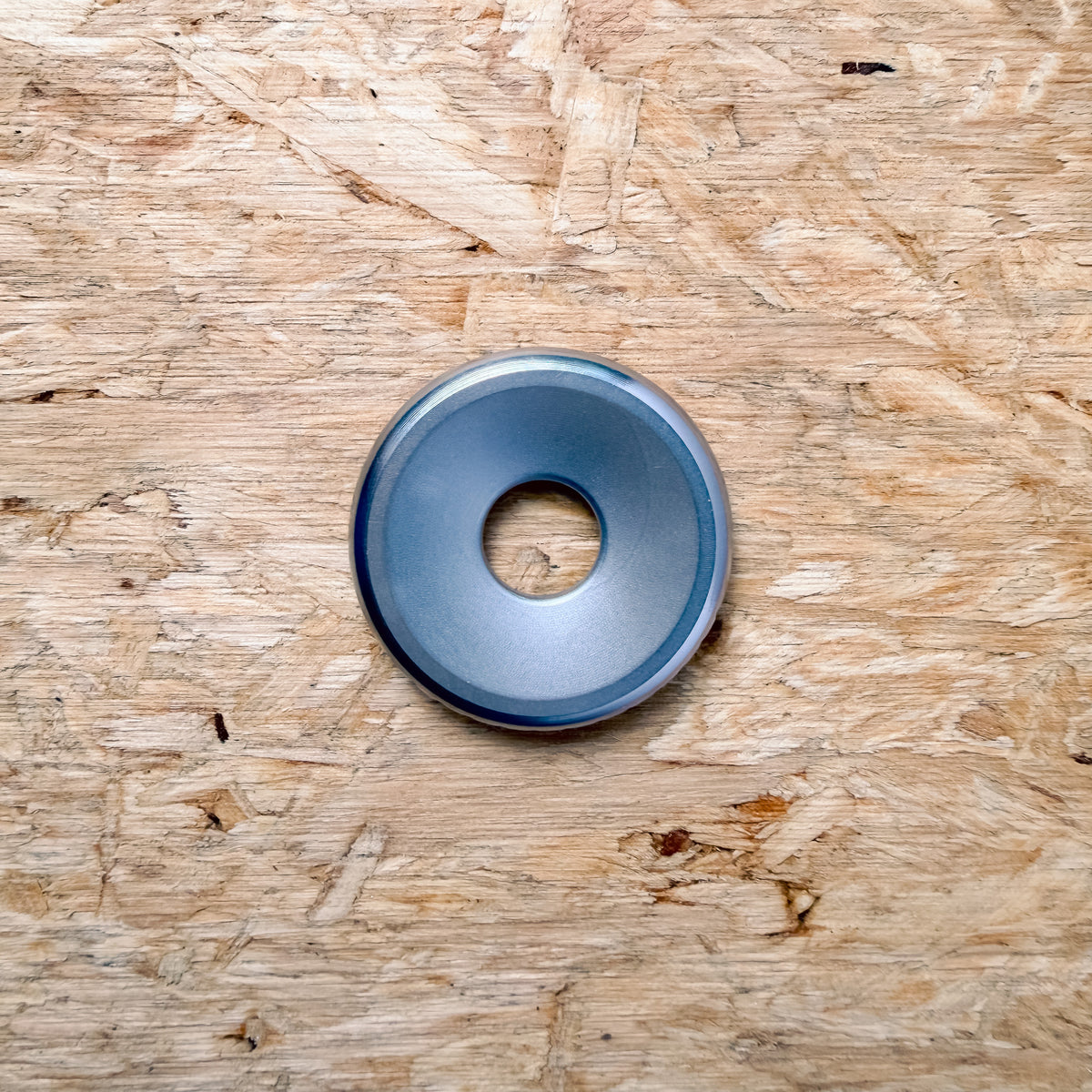 A circular metallic 316 stainless steel ring or coin with a smooth surface, placed flat on a textured plywood or OSB surface.