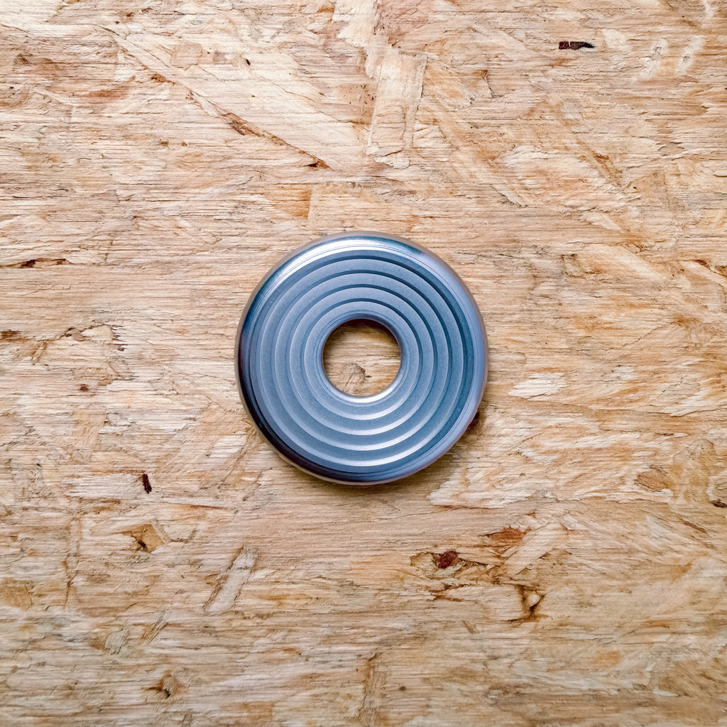 A circular metallic 316 stainless steel ring or coin with concentric grooves, placed flat on a textured plywood or OSB surface.