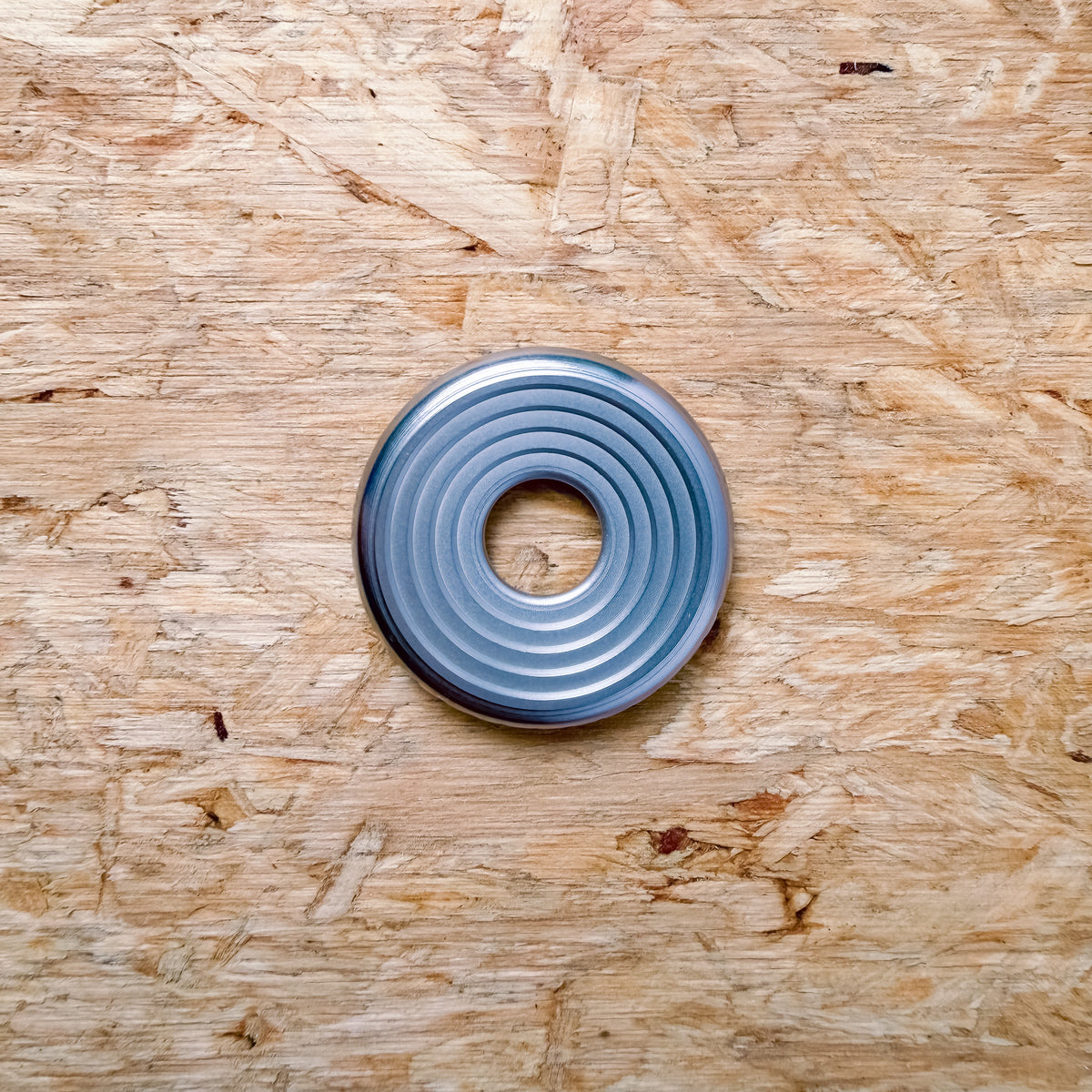A circular metallic 316 stainless steel ring or coin with concentric grooves, placed flat on a textured plywood or OSB surface.