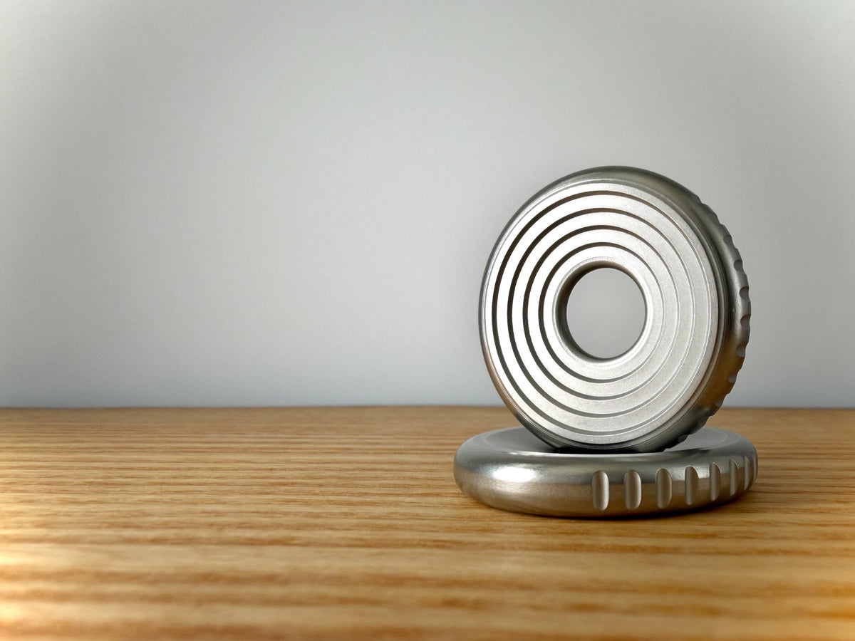 A pair of metallic silver 316 stainless steel circular coin-shaped objects, one standing upright and one lying flat, displayed on a light Ash wood surface against a neutral gray background.