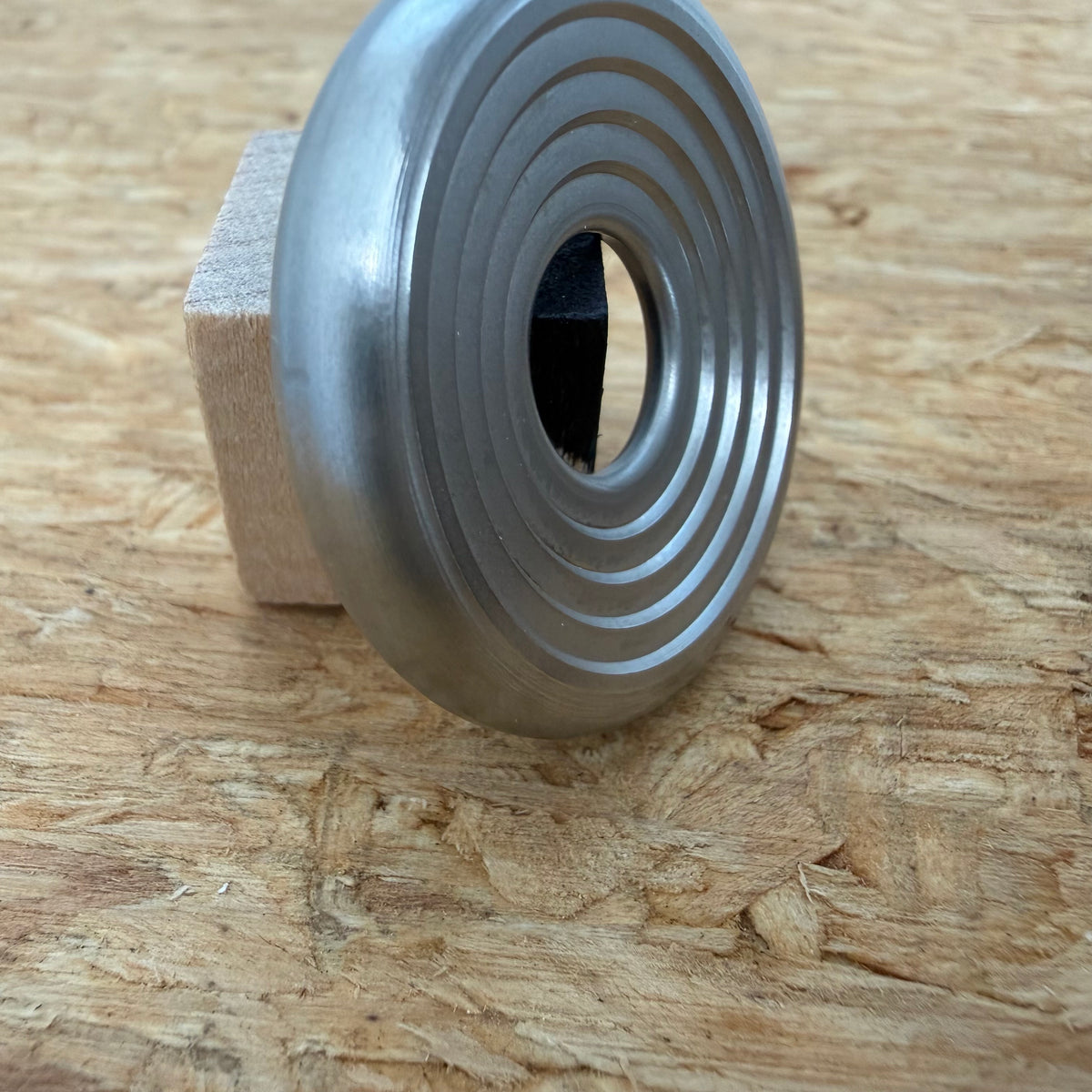 A 316 stainless coin-shaped object with concentric grooves and a hollow center, resting against a small wooden block on a textured wood surface.