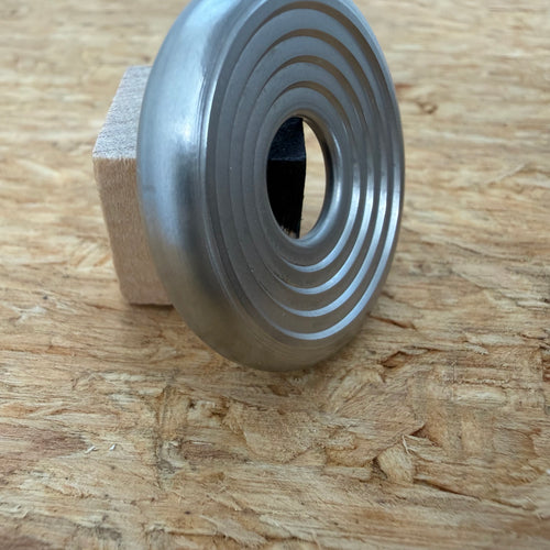 A 316 stainless coin-shaped object with concentric grooves and a hollow center, resting against a small wooden block on a textured wood surface.