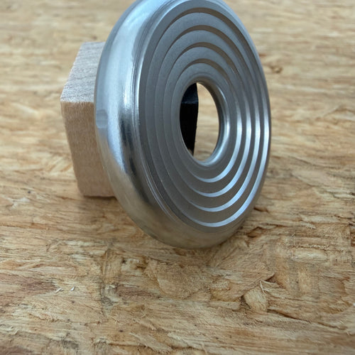 A 316 stainless coin-shaped object with concentric grooves and a hollow center, resting against a small wooden block on a textured wood surface.