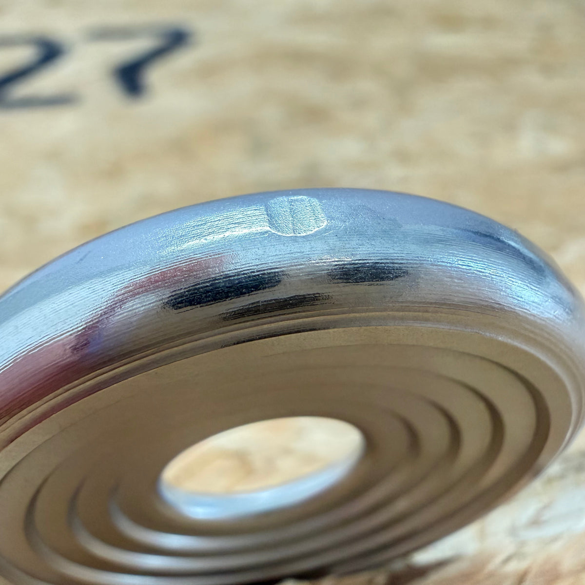 A close up of a 316 stainless coin-shaped object with concentric grooves and a hollow center, set against a blurred wood backdrop.
