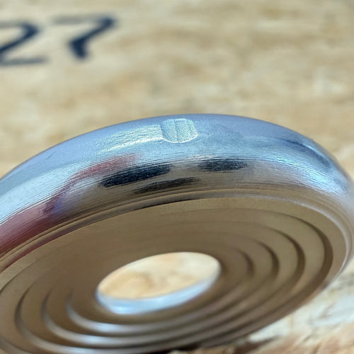 A close up of a 316 stainless coin-shaped object with concentric grooves and a hollow center, set against a blurred wood backdrop.