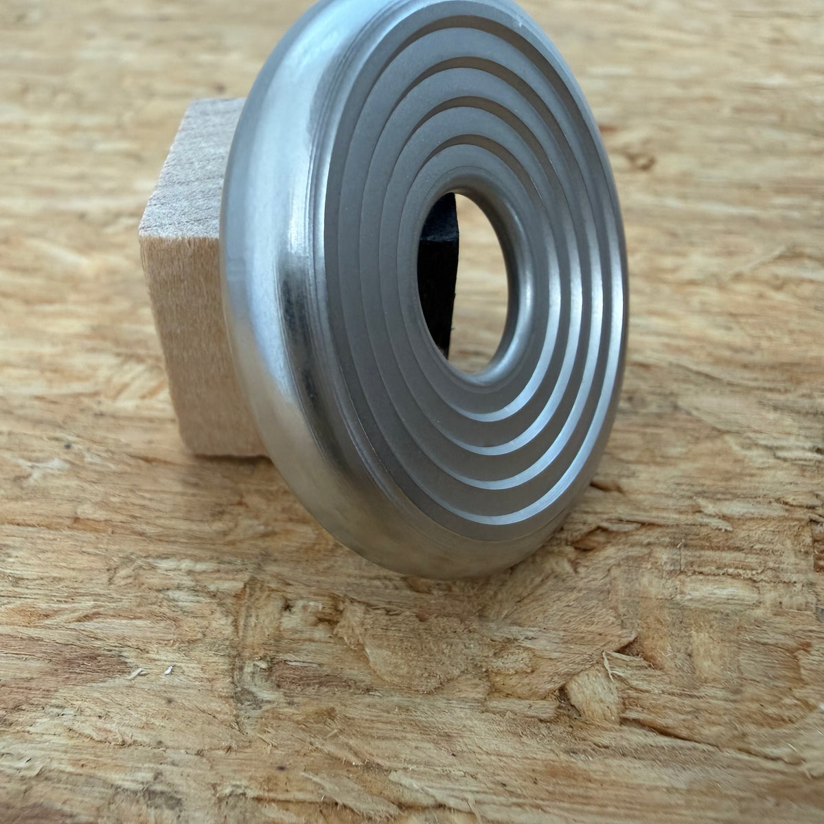 A 316 stainless coin-shaped object with concentric grooves and a hollow center, resting against a small wooden block on a textured wood surface.