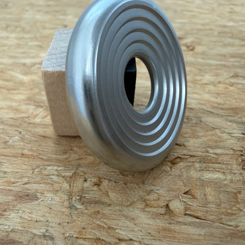 A 316 stainless coin-shaped object with concentric grooves and a hollow center, resting against a small wooden block on a textured wood surface.
