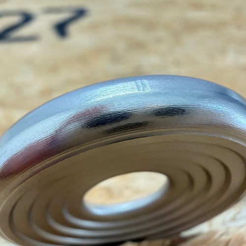 A close up of a 316 stainless coin-shaped object with concentric grooves and a hollow center, set against a blurred wood backdrop.