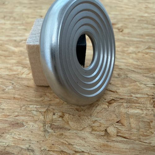A 316 stainless coin-shaped object with concentric grooves and a hollow center, resting against a small wooden block on a textured wood surface.