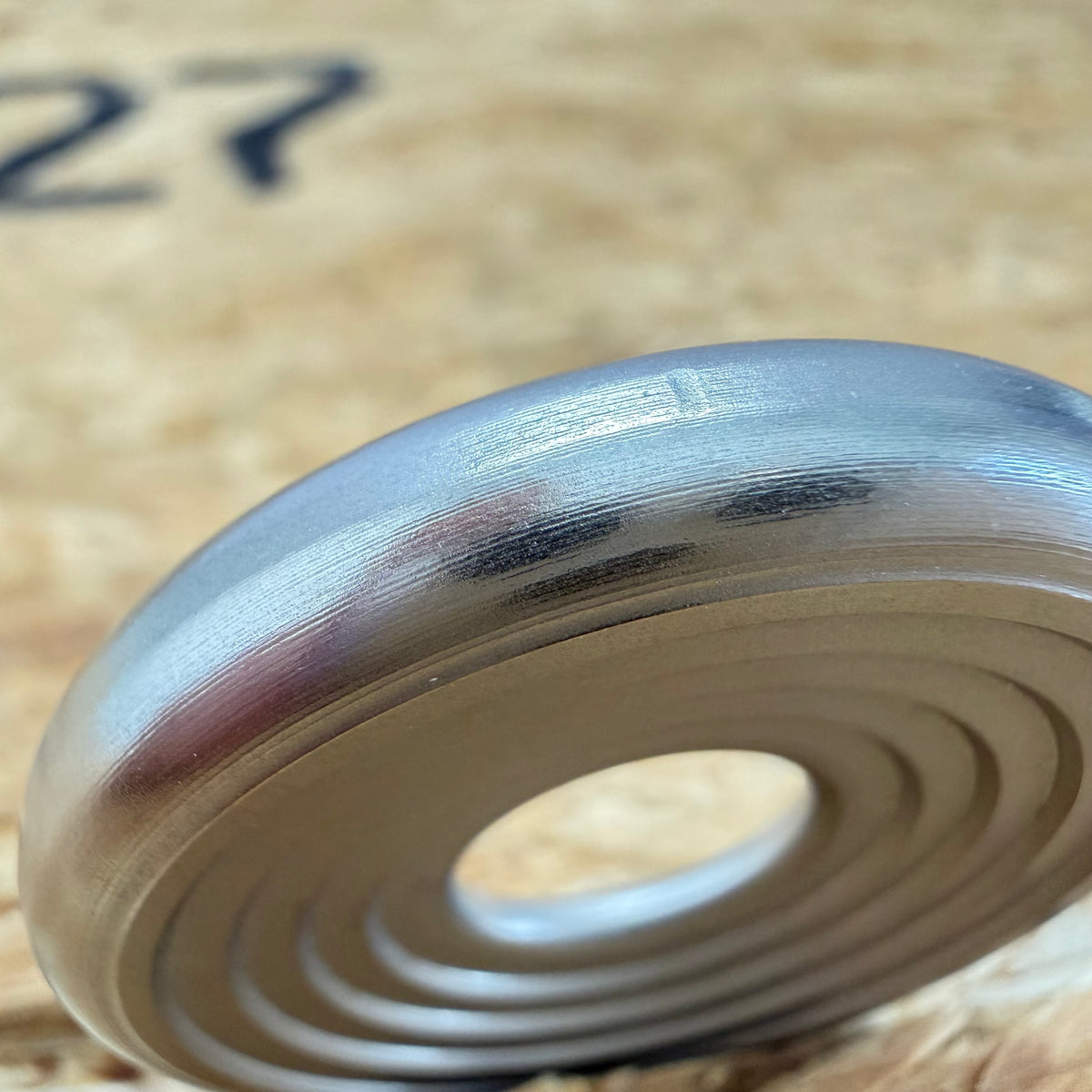 A close up of a 316 stainless coin-shaped object with concentric grooves and a hollow center, set against a blurred wood backdrop.