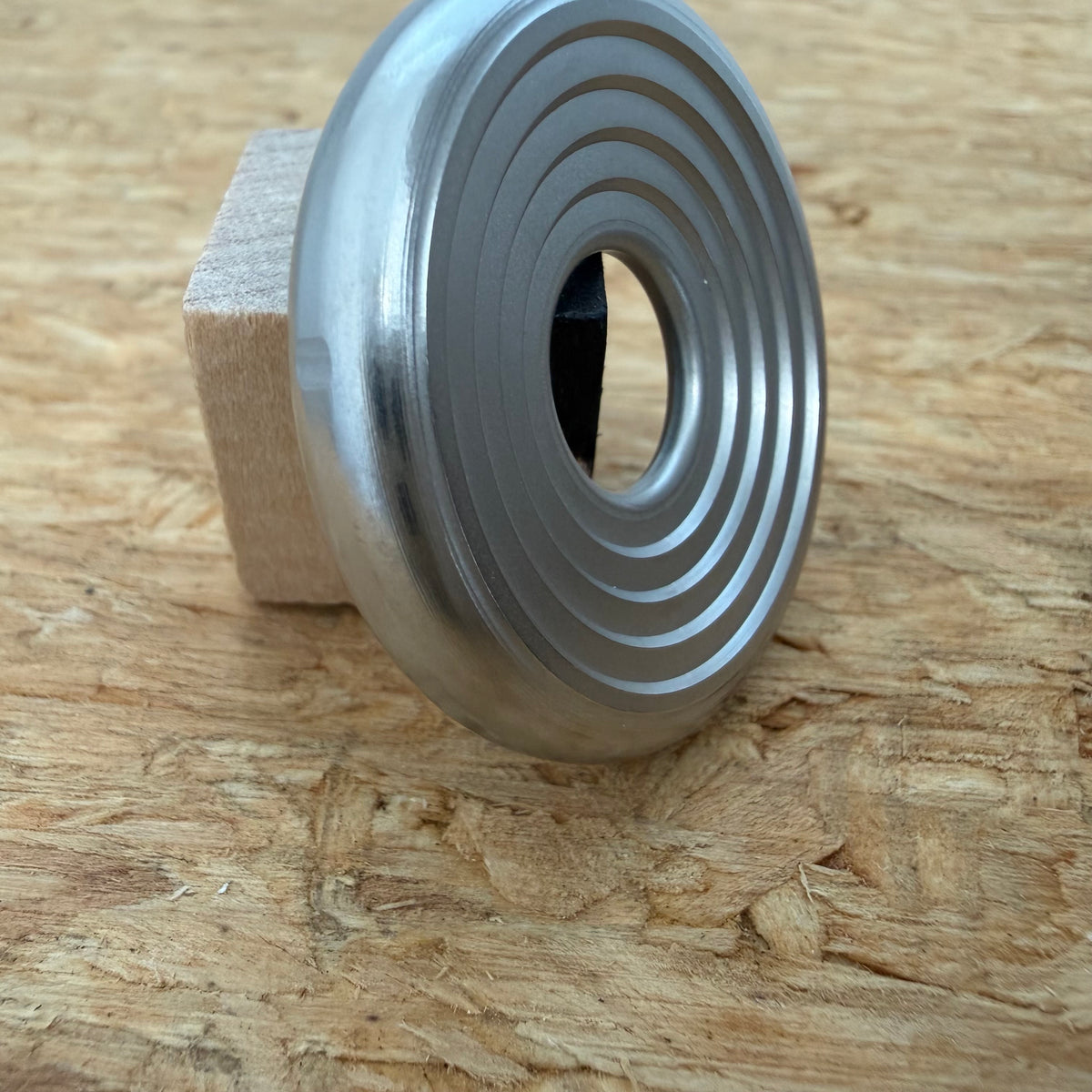 A 316 stainless coin-shaped object with concentric grooves and a hollow center, resting against a small wooden block on a textured wood surface.