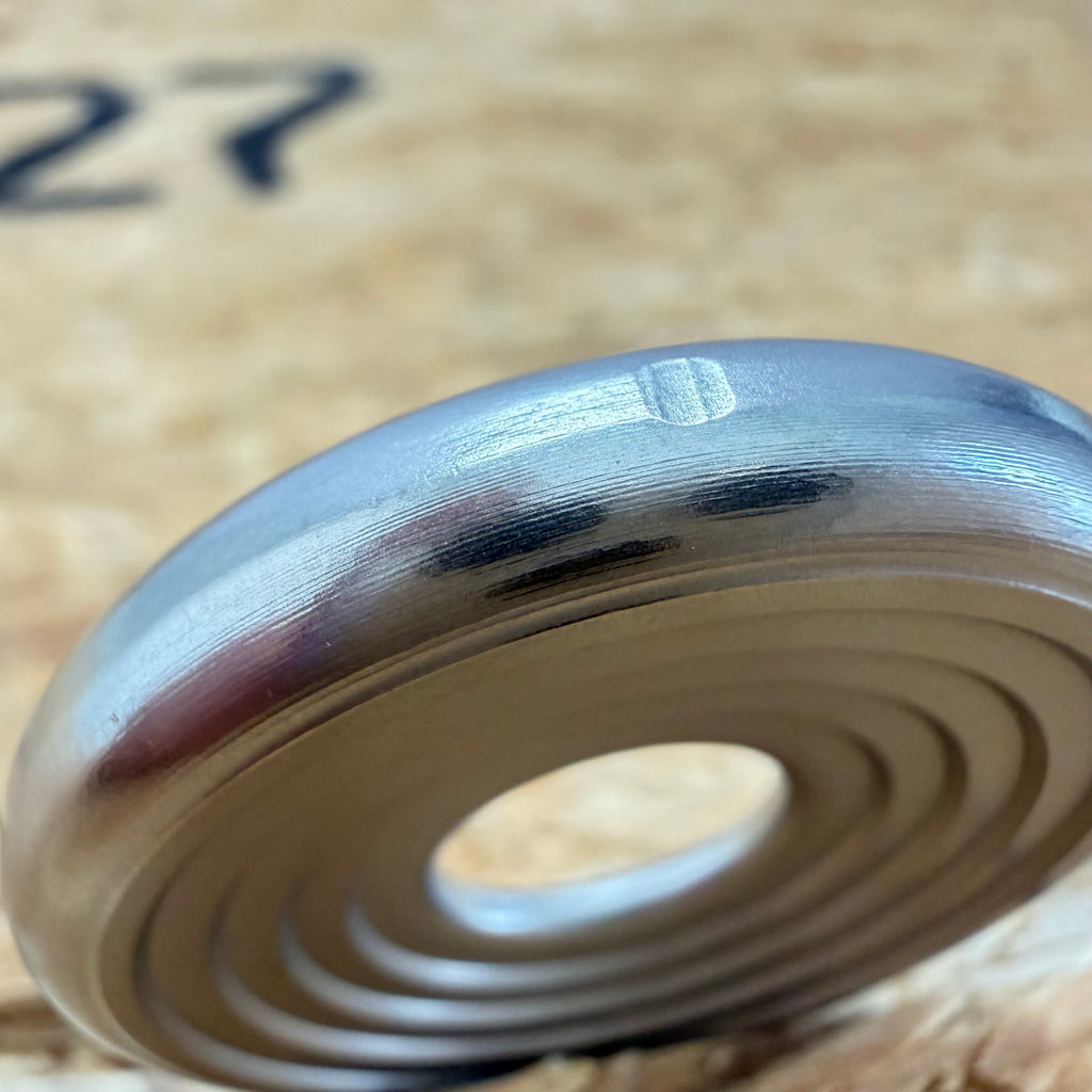 A close up of a 316 stainless coin-shaped object with concentric grooves and a hollow center, set against a blurred wood backdrop.