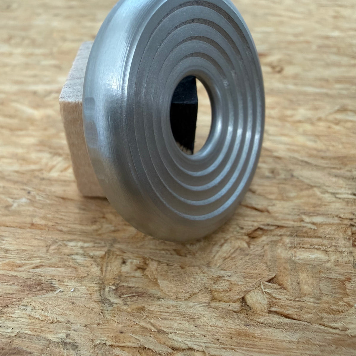 A 316 stainless coin-shaped object with concentric grooves and a hollow center, resting against a small wooden block on a textured wood surface.