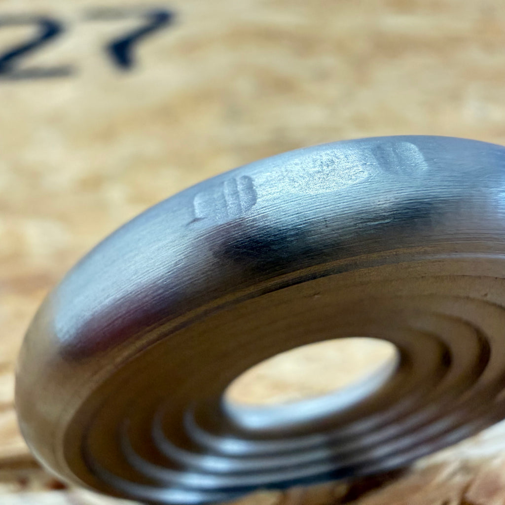 A close up of a 316 stainless coin-shaped object with concentric grooves and a hollow center, set against a blurred wood backdrop.
