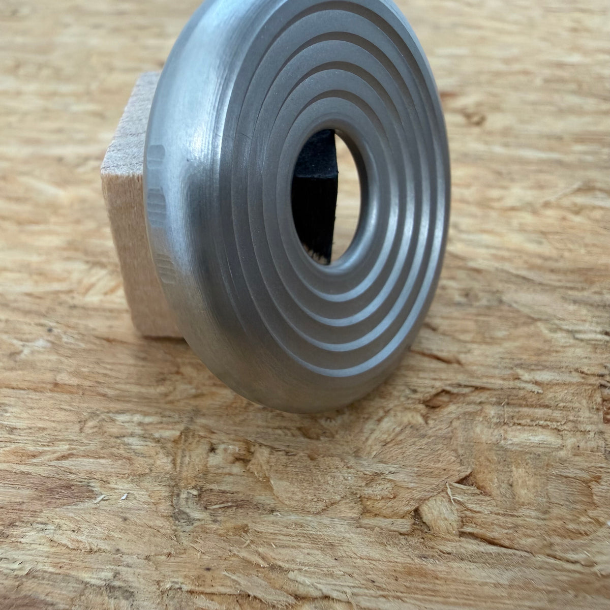 A 316 stainless coin-shaped object with concentric grooves and a hollow center, resting against a small wooden block on a textured wood surface.