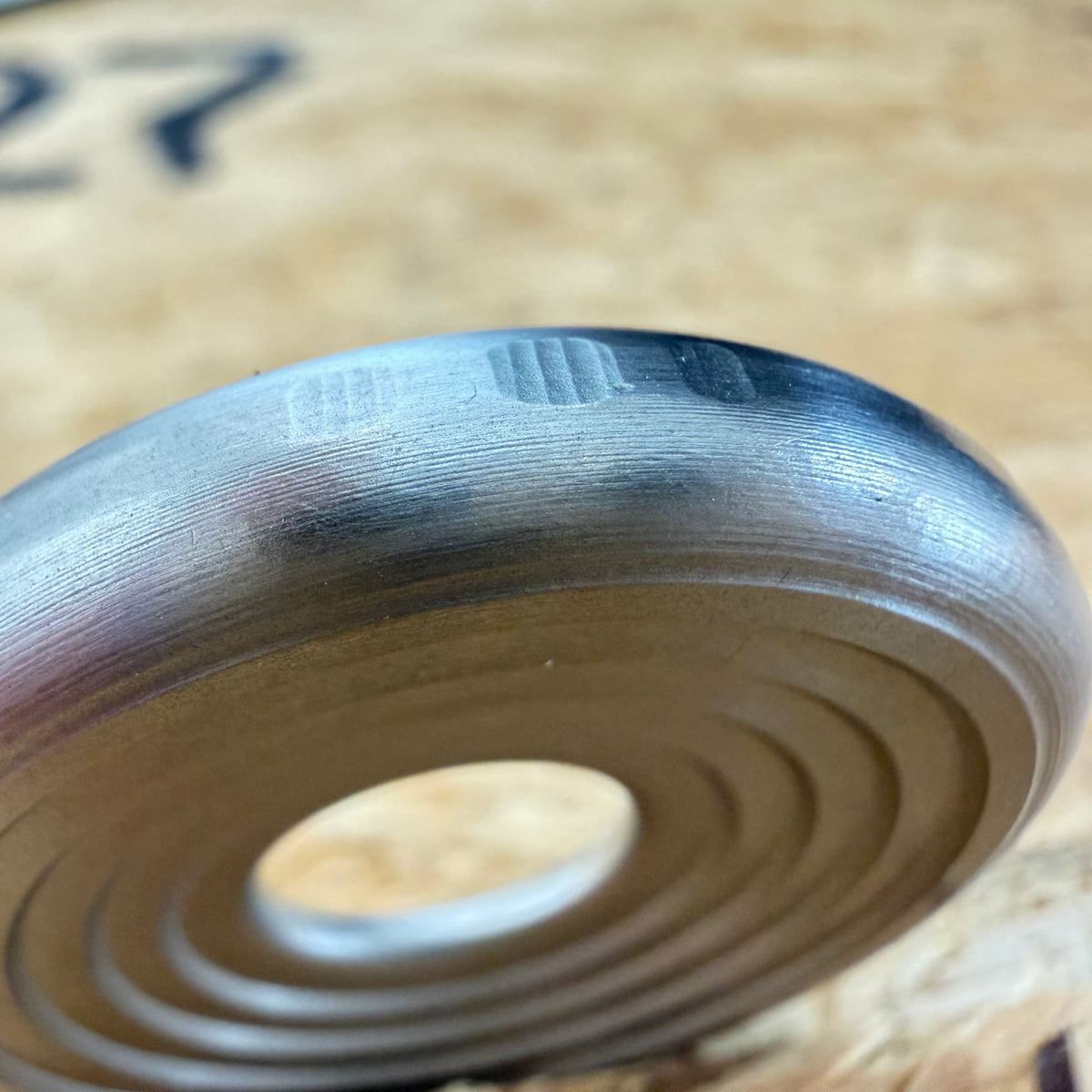 A close up of a 316 stainless coin-shaped object with concentric grooves and a hollow center, set against a blurred wood backdrop.